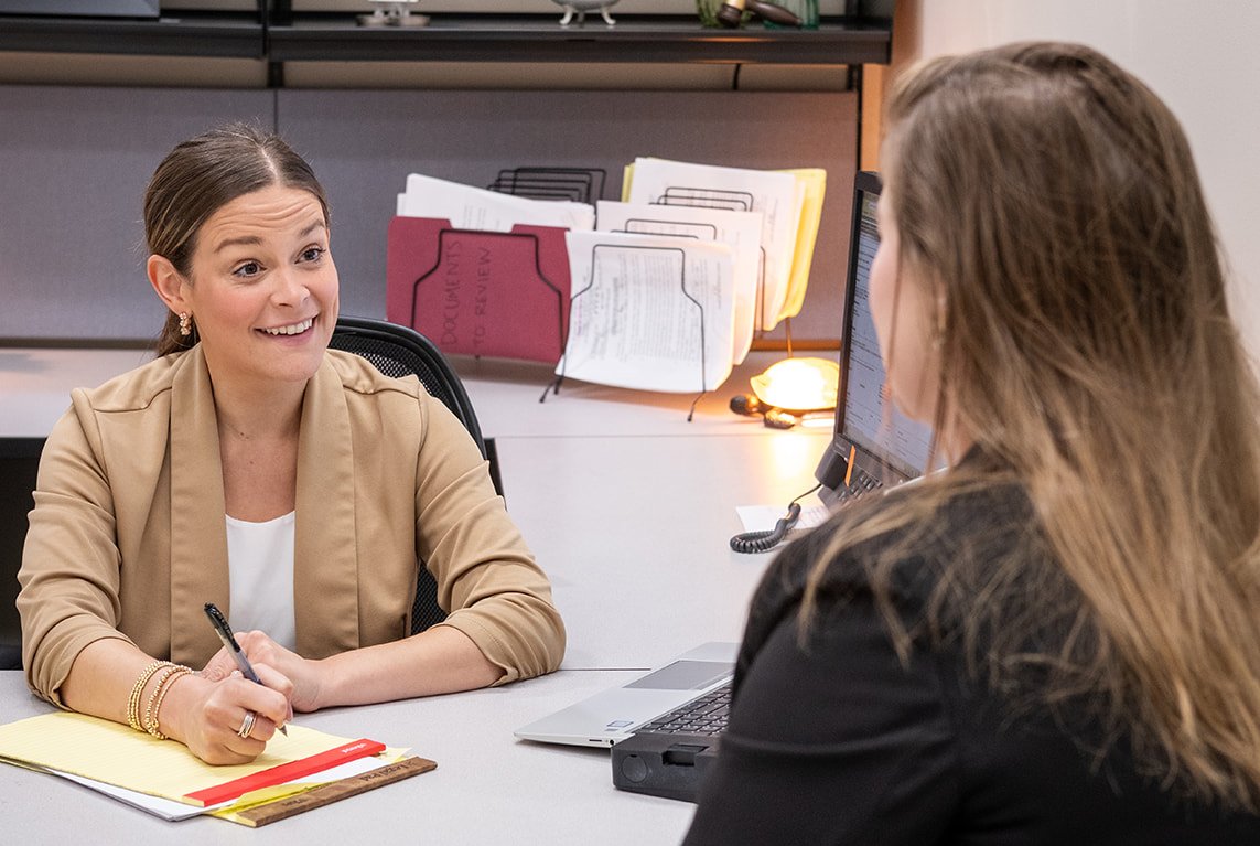 Woman behind desk facing the camera, taking notes while conversing with woman on other side of desk